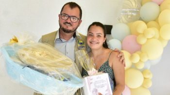 Secretário municipal posa ao lado de uma gestante durante a entrega de kit de natalidade promovida pela Prefeitura de Correntina. A gestante segura um certificado e itens do kit enquanto ambos sorriem diante de uma decoração com balões.