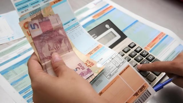 Pessoa segurando cédulas de dinheiro em uma mão enquanto outra mão utilizando uma calculadora, com contas de energia elétrica e faturas espalhadas sobre a mesa.