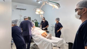 Sala de cirurgia em unidade de saúde, com equipe médica utilizando equipamentos hospitalares durante procedimento cirúrgico em paciente deitado na mesa operatória.