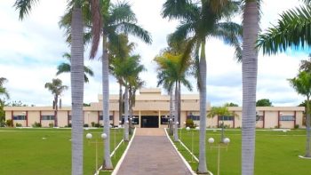 Fachada de um prédio público de um andar de cor bege, antecedido por um grande gramado verde e um caminho de pedras ladeado por palmeiras imperiais altas e postes de iluminação de jardim sob um céu parcialmente nublado.