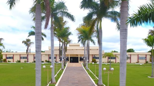 Fachada de um prédio público de um andar de cor bege, antecedido por um grande gramado verde e um caminho de pedras ladeado por palmeiras imperiais altas e postes de iluminação de jardim sob um céu parcialmente nublado.