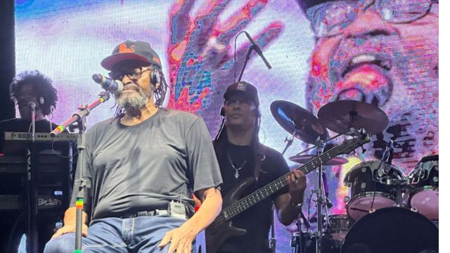 Cantor de reggae se apresenta no palco, sentado em frente ao microfone, acompanhado por banda com baixo, bateria e teclado, durante show noturno com telão iluminado ao fundo.