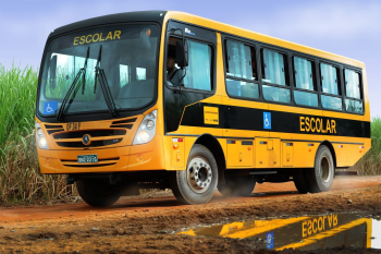 Ônibus escolar amarelo e preto circulando por estrada de terra na zona rural, com placa de acessibilidade visível e estudantes no interior do veículo. Ao fundo, há vegetação de canavial sob céu claro.