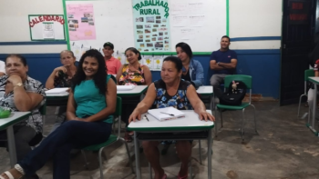 Alunos adultos e idosos participam de aula da Educação de Jovens, Adultos e Idosos (EJAI) em sala de aula em Correntina. Sentados em carteiras escolares, alguns sorriem enquanto acompanham a aula com cadernos e canetas sobre a mesa. Ao fundo, há cartazes coloridos com o tema 'Trabalhador Rural' e um calendário na parede.