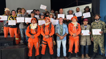 Grupo de novos brigadistas voluntários de Correntina posa para foto oficial após a conclusão do Curso de Combate a Incêndios Florestais. Na imagem, 19 formandos seguram seus certificados ao lado de instrutores do Corpo de Bombeiros de Barreiras, todos uniformizados com trajes de proteção. A cerimônia marca o encerramento do curso promovido pela Prefeitura de Correntina e Secretaria Municipal de Meio Ambiente, com foco na prevenção e resposta a incêndios na zona rural. A imagem foi registrada em ambiente fechado com todos sorrindo e celebrando a formação.